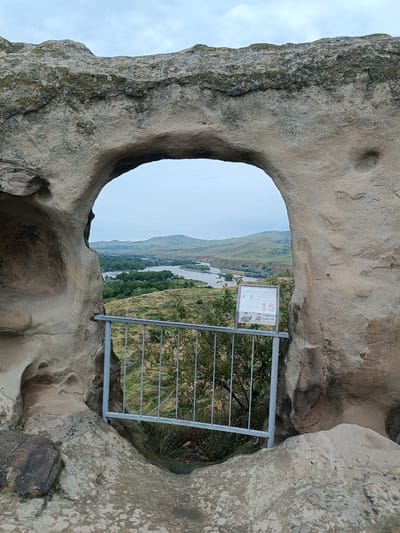 The image shows a natural rock archway in the ancient cave city of Uplistsikhe, Georgia. Through the arch, a scenic view of the Mtkvari River and surrounding rolling hills unfolds under an overcast sky. A metal railing and a small informational sign are visible in the foreground, positioned just outside the arch. The rock formations are weathered and textured, indicating their age. No people are present in the scene. The environment is natural and historical, characterized by rock structures, vegetation, and a river valley.