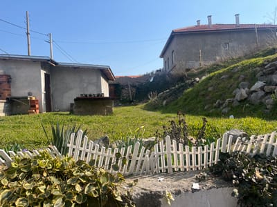 The image depicts an outdoor residential or garden scene in Vetren, Bulgaria, under a clear blue sky. In the foreground, a low, white picket fence section rests on a concrete ledge, with variegated green and yellow foliage directly in front of it. Behind the fence, a grassy lawn extends into the midground. To the left, a single-story, light-grey building with a dark wooden door is visible. Adjacent to its left side is a brick structure, possibly an outdoor oven or grill, with a stack of red bricks nearby. To the right of the single-story building, a concrete block structure with a dark metal grate or wooden slats on top stands on the grass. Two utility poles with overhead wires are present in the upper-left portion of the frame. To the right, a grassy hillside, interspersed with grey rocks, slopes upward. Atop this slope, a larger, two-story building with a light-colored exterior, a red-tiled roof, and three prominent grey chimneys is positioned. No individuals or specific actions are depicted in the scene.