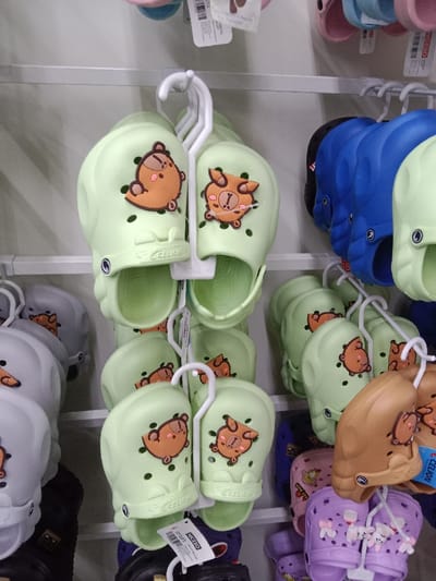 This image depicts a retail display of numerous pairs of children's closed-toe clog-style footwear, consistent with a store environment in Kauman, Indonesia. The footwear is organized on white metal shelving units.
The primary subjects are pairs of shoes, predominantly light green, gray, blue, black, brown/tan, pink, and purple. Each pair features a decorative charm affixed to the upper section of the shoe. Many of the light green and gray pairs display a brown, cartoonish animal character, possibly a bear or a similar creature, with variations in expression (e.g., sleeping or awake). The word "CELION" is embossed on the side of many shoes. A purple pair at the bottom right includes multiple white, three-dimensional charms resembling small animals, possibly bunnies, with bows.
Each pair of shoes is suspended from a white plastic hanger. A white product tag, partially obscured, with "NICESO" visible, hangs from one pair. The background consists of more shelving and indistinct store elements, indicating an indoor retail setting. No individuals are present in the frame.