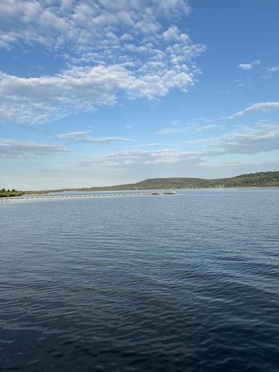 A wide shot shows a body of water with calm ripples. In the distance, a long, white structure stretches across the water, appearing to be a pier or a barrier. Beyond the water, a line of trees and hills forms the horizon under a blue sky dotted with white, puffy clouds. The scene is tranquil and serene, with no people or discernible activity. The location is identified as Kazashko, Bulgaria.