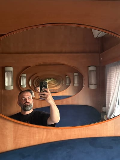 A man with a beard is taking a mirror selfie in a room with wood paneling. The room has a curved wooden wall with several oval mirrors that create an infinite reflection effect. There are light fixtures on the wall. The man is wearing a black t-shirt and is holding a smartphone in his right hand. The floor is covered with a blue fabric. The scene appears to be in a residential or recreational setting. There is no visible indication of the location being in Mariinsk, Russia.