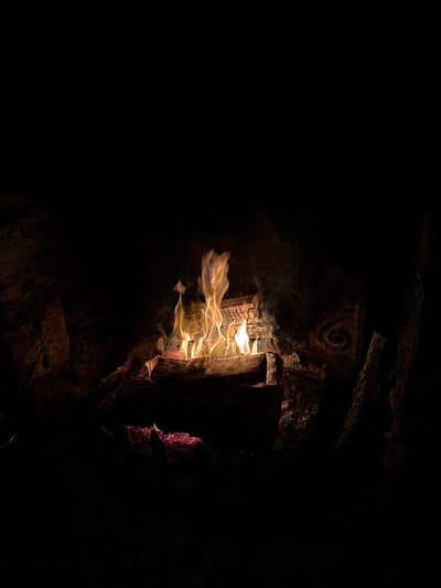 A dark, cavernous space with a structure built of rocks and wood. Firelight flickers around the wood, creating long streaks of golden light. A light smoke rises from the structure, fading into the darkness. An etched spiral symbol is visible on the rock above the structure. The overall scene is dimly lit, emphasizing shadows and creating a sense of mystery.