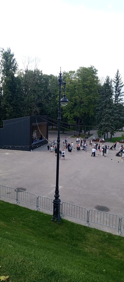 A high-angle shot captures an outdoor public space, likely a park or plaza, with a stage set up on the left. The stage is a modern, dark-colored structure with a sloped roof and vertical siding. A few individuals are visible on the stage, one appearing to be a performer with a microphone. Strings of small lights are draped across the stage and extend into the surrounding area.

The ground is paved with light-colored interlocking bricks. A prominent black lamppost with an ornate design stands in the foreground, slightly off-center to the right, with a clear glass lantern. In the middle ground, a crowd of people are gathered, some standing, some sitting on benches or on the ground. They are dressed in casual attire. Several strollers and a baby stroller are also present.

Beyond the paved area, lush green grass can be seen, bordered by a silver metal fence. In the background, a dense line of mature trees forms a natural backdrop, with a few evergreen trees interspersed. The sky above is bright and appears overcast. The scene suggests a public event or gathering is taking place. The location is identified as Dub'e, Russia.
