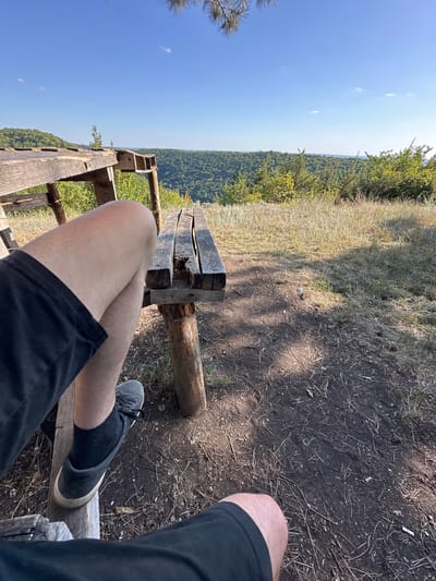 The image captures a person's legs and feet, clad in black shorts and dark socks with grey sneakers, resting on a wooden bench. The bench appears weathered and rustic, with visible grain and some signs of wear and tear. The setting is outdoors, likely a park or scenic overlook, with a panoramic view of a lush, green, forested valley stretching into the distance under a clear blue sky. The ground around the bench is a mixture of dry grass, dirt, and scattered pine needles. The lighting suggests it is daytime, possibly late afternoon, given the dappled shadows cast on the ground. The scene evokes a sense of relaxation and appreciation of nature. The specific location within Velykozalissia, Ukraine, cannot be determined solely from the visual information provided.