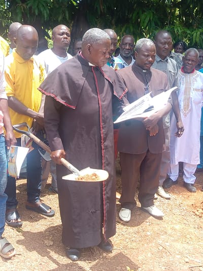 A group of people stand in a dirt clearing in Ninzo South, Nigeria. A man in a black robe with red trim holds a shovel with dirt on it. Another man in a brown suit holds a document. Other people surround them, some in casual clothing and others in more formal attire. A tree is visible in the background.