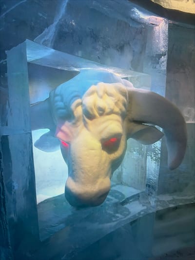 The image displays a detailed ice sculpture of a bull or ox head, positioned within an ice-built structure in Kiruna, Sweden. The sculpture is rendered in white and features prominent, upward-curving horns. Its eyes are distinctly illuminated with a red light. The surrounding environment consists of large, translucent blocks of ice forming walls and structural elements, giving the scene a cool, blue-toned ambiance. The sculpture is the central subject, static and imposing. No people, active movements, or direct interactions are visible.