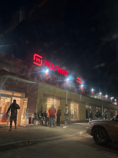 The image depicts an evening scene in Сурок, Russia, outside a supermarket. The most prominent feature is the store's large, illuminated red sign in Cyrillic script, displaying "Магнит" (Magnit), a well-known Russian supermarket chain. Below it, another red sign reads "КЕЙВЫТ МАГАЗИН," likely indicating a convenience store or a specific type of shop within the complex. The building's facade consists of light-colored brickwork on the lower section and a darker, patterned cladding above, beneath the main sign.
Several people are gathered outside the store. In the immediate foreground, a person in dark attire is walking near the brightly lit glass entrance, holding an object. Further down the sidewalk, towards the left of the entrance, a small group of individuals are standing, some appearing to be waiting or socializing. One person among them is wearing a backpack. Closer to a parked car, another small group of people are standing and conversing.
A light-colored sedan or hatchback is parked on the street next to the building. The scene is set at night, with a dark sky and the entire area illuminated by the store's bright signage, interior lights, and additional exterior spotlights, which cast reflections on the ground, suggesting it might be wet or have a reflective surface. The visible activity consists of people moving and congregating outside the establishment, consistent with a typical evening at a local market.