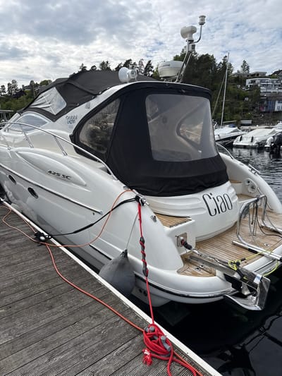 A partially-covered white boat is docked at a wooden pier. The boat is tied to the pier with a red rope. The name "CIRO" is visible on the boat's rear. "425 SC" is printed on the side of the boat, along with "Gobb LH2781." Gray fenders are between the boat and the pier. Other boats are visible in the background, moored in the water. Buildings and trees are visible on a hill in the background. The photo was taken in Nesøya, Norway. The sky is cloudy.