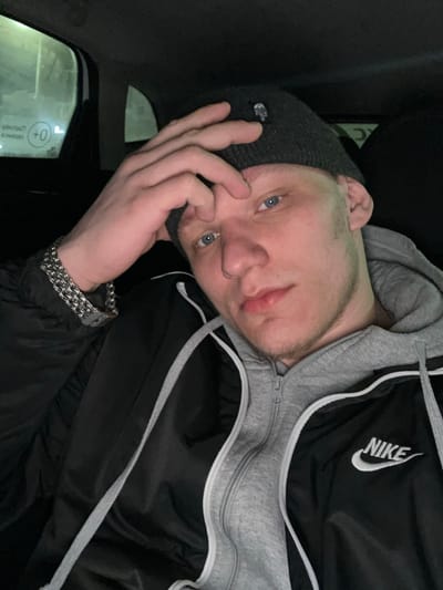 The image depicts a young adult male inside a vehicle, located in Dobryanka, Russia. The male, with light skin and blue eyes, displays light stubble on his chin and jawline. He is wearing a dark grey knitted beanie adorned with a small white skull logo, a black jacket featuring a white "NIKE" logo on the right chest, and a grey hooded sweatshirt worn underneath. A multi-strand silver-colored chain bracelet is visible on his left wrist. His right hand is raised, with fingers resting on his forehead and beanie. He is looking directly forward, engaging with the camera. The vehicle's interior is predominantly dark. Through the rear passenger window on the left, a blurry, light-colored exterior or reflection is visible, along with inverted and indistinct Cyrillic text that appears to read "CPEA +0" or similar.
