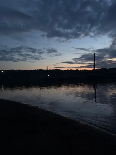 This image depicts a river scene at dusk in the city of Zaozer'e (Khokhlovskoe S/P), Russia. The sky is filled with dramatic, dark clouds, with a hint of orange and blue from the setting sun visible on the horizon. A tall, industrial smokestack stands silhouetted against the sky on the far bank, which is densely wooded. Lights can be seen dotting the treeline, suggesting some form of settlement or industrial activity. The river's surface is calm, reflecting the dim light and the dark silhouette of the opposite shore. In the foreground, a dark, sandy bank slopes down to the water, partially obscuring the reflection. There are no people visible in the frame. The overall atmosphere is somber and tranquil.