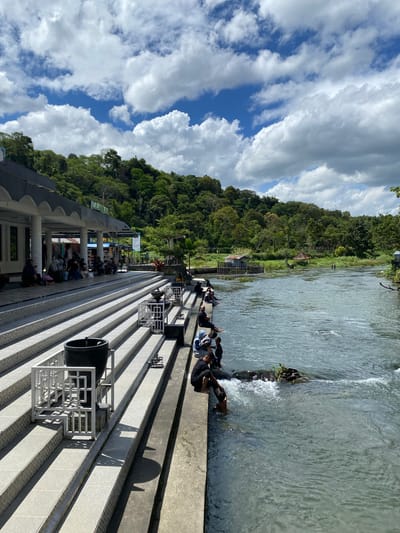 In Blang Bebangka, Indonesia, a wide, flowing river runs alongside a tiered, white-tiled staircase structure. The staircase is bordered by low white metal railings and leads up to a covered building with an arched roofline, where several people are seated on the tiled floor. More individuals are positioned on the steps of the staircase, some with their feet in the river. Further down the riverbank, several people are wading and playing in the water. The river is clear with visible currents and a few scattered rocks, some with vegetation. In the background, a lush green, tree-covered hillside rises up, meeting a bright blue sky dotted with fluffy white clouds. Small, rustic structures with thatched roofs are visible along the riverbank. The overall scene depicts a serene, recreational setting on a sunny day.