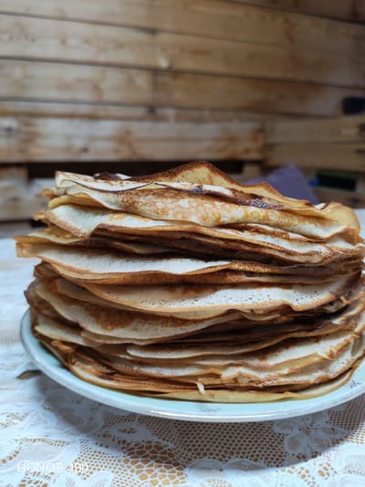 A close-up shot focuses on a tall stack of thin, golden-brown pancakes, or blini, on a light blue plate. The pancakes are layered unevenly, creating a textured and inviting appearance. Some edges show darker, slightly crisped brown spots from cooking. The plate rests on a white lace tablecloth with an intricate floral pattern, which is itself laid over a textured brown surface.
In the background, a wooden plank wall creates a rustic setting. The wood appears to be aged, with visible grain and horizontal planks. The lighting suggests it might be daytime indoors, with a soft light illuminating the pancakes and casting subtle shadows. There are hints of other objects in the blurred background, including what appears to be a dark rectangular shape and a muted purple fabric.
The scene evokes a sense of home cooking and comfort, suggesting a meal being prepared or served. The focus on the freshly made pancakes implies a recent activity of baking. No people are visible.
The visible text at the bottom of the image reads "HONOR 400". The location context is identified as Эрхирик, Russia.