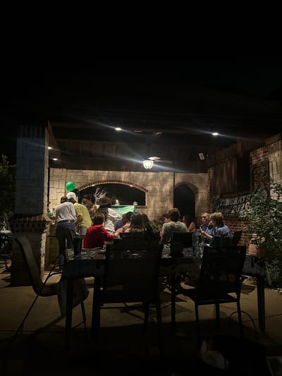 The image shows an outdoor gathering at night, likely in a backyard setting in Southlake, Texas, USA. A group of people are seated around tables, seemingly engaged in a meal or activity. The lighting is dim, with visible light fixtures illuminating the area. Stone and brick structures form the backdrop. A banner or sign that says "Congrats Class of 2026" can be seen. There are also some balloons. There are no obvious signs of distress or unusual activity.