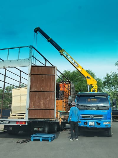 In Boralday, Kazakhstan, a blue flatbed truck with a crane is parked on an asphalt surface. The crane, which is yellow and black, is extending over the truck's bed, which is filled with wooden crates. A person wearing an orange shirt stands on top of the crates in the truck bed. Another person, wearing a blue long-sleeved shirt and dark pants, stands behind the truck, facing away from the camera. Two blue plastic pallets are stacked on the ground next to the truck, and a metal tool is also on the ground. The sky is bright blue, and trees are visible in the background. The license plate on the front of the truck reads "KZ 257 16 AEY". The truck has the brand name "DFAC" visible on the front.