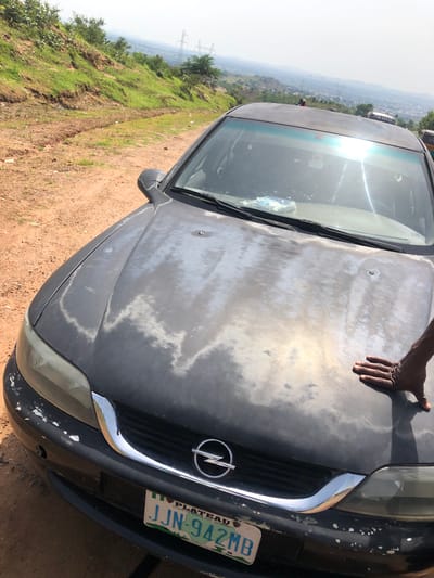 A close-up, high-angle shot shows the heavily weathered hood of a dark-colored Opel car parked on a dirt road. The paint on the hood is peeling and faded, with lighter patches showing through. A license plate on the front bumper reads "JJN 942MB" and displays the text "PLATEAU HOME OF PEACE AND TOURISM." To the right of the car, a dark-skinned hand is resting on the hood. In the background, a green, grassy hillside slopes upwards, with power lines and transmission towers visible on the horizon. The sky is bright and hazy. The scene appears to be an outdoor, possibly rural or semi-urban, location in Plateau State, Nigeria.