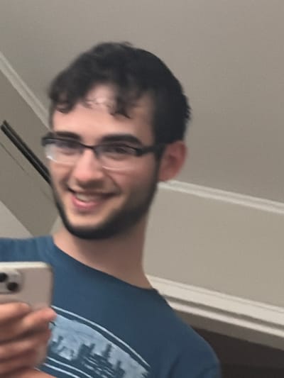 The image captures a young man taking a selfie indoors in Franklin Township, United States of America. The photograph is rotated 90 degrees clockwise. The subject is a man with dark, curly hair, a beard, and glasses, smiling broadly at the camera. He is wearing a blue t-shirt with a light-colored graphic design, which appears to depict a skyline or abstract pattern on the chest area. His left hand is visible in the bottom right corner, holding a smartphone (likely an iPhone, based on the visible camera module), which he is using to take the picture. The background consists of a plain, light-colored wall and a vertical white door frame or trim, suggesting a typical indoor residential setting. There is no visible text in the image. The man's joyful expression indicates a positive mood.