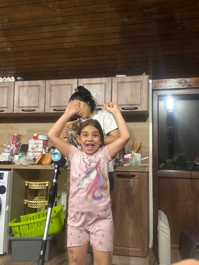 In what appears to be a kitchen area in Ruska Bela, Bulgaria, two individuals are present. A young girl in the foreground, wearing pink pajamas with a unicorn print, is excitedly raising her arms and has a wide, open-mouthed expression. Behind her, a woman, possibly her mother, is partially visible, holding what appears to be a small pet and raising one hand. The kitchen features wooden cabinets above and below the countertop, a sink area with cleaning supplies, and a white washing machine to the left. In front of the girl stands a black and silver vacuum cleaner. The overall impression is one of a domestic scene, possibly during playtime or a moment of energetic activity within the home.