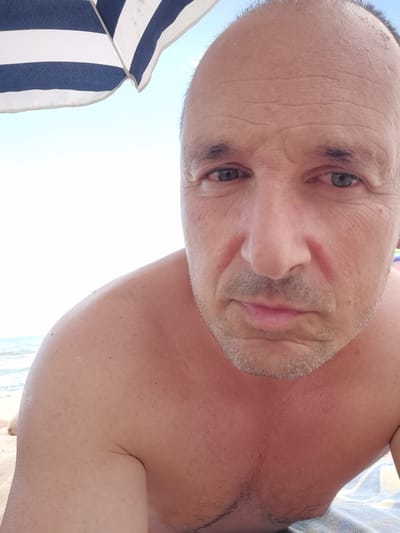 The media file is a close-up, low-angle selfie of a middle-aged man at the beach in Osenovo, Bulgaria. He is shirtless, with his upper chest and shoulders visible. His skin appears slightly reddened, suggesting he may have a sunburn. A portion of a blue and white striped beach umbrella is visible in the upper left corner of the frame. The background shows the ocean and a bright, clear sky, indicating it is daytime and the weather is sunny. The man has short hair and stubble on his chin and upper lip. His expression is neutral. There is no visible text in the media file.