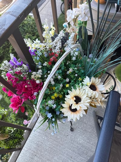 The image depicts a close-up, high-angle view of a vibrant artificial flower arrangement in a white woven basket. The basket is filled with a variety of flowers and greenery, including large white sunflowers with dark centers, bright pink peonies, purple bell-shaped flowers, small red and white clusters, and delicate blue blossoms. Green foliage and slender, light green ornamental grasses also contribute to the fullness of the arrangement. The basket is placed on a concrete surface, likely a balcony or patio, next to a dark gray metal railing. In the background, glimpses of lush green trees and other foliage are visible. A portion of a dark gray outdoor chair is visible on the right side of the frame. The scene is well-lit, suggesting it was taken outdoors during the daytime. There is no indication of Philadelphia, United States of America.