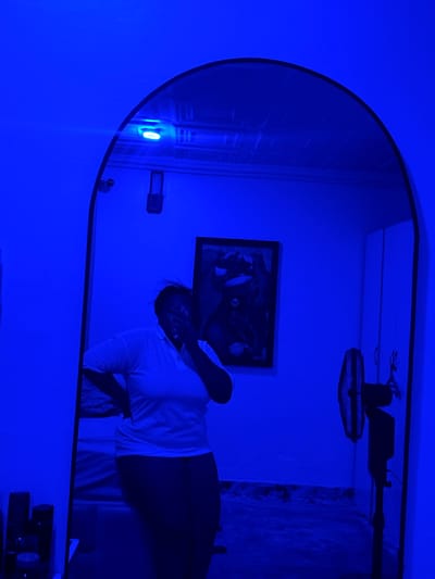The image depicts a mirror's reflection in Rumuigbo, Nigeria. The reflection shows a woman in a white shirt, a picture frame on the wall, a ceiling fan, and what appears to be weights in the lower right. The room is illuminated with a strong blue light. The mirror itself has a rounded edge.