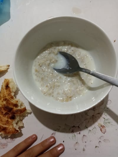 An overhead shot captures a white bowl filled with oatmeal and milk, a spoon resting inside, positioned on a light-colored tablecloth with a floral pattern. To the left of the bowl are two pieces of torn flatbread, one appearing toasted. In the lower portion of the image, a person's hand with manicured fingernails is visible, suggesting a meal is being consumed. The scene is indoors, likely a kitchen or dining area. The image does not provide specific visual cues to determine the location within the city of Кош-Дөбө, Kyrgyzstan.