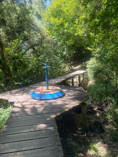 The image shows a colorful, circular merry-go-round situated on a wooden platform in a forest setting. The merry-go-round is blue with a brightly colored disc featuring patterns of red, yellow, and blue. A blue pole extends vertically from the center of the disc. The wooden platform is made of planks and appears to be part of a walkway or bridge in a wooded area. In the background, a second wooden bridge crosses over a small stream or ditch, leading uphill into denser foliage. The surrounding environment is lush and green, with sunlight filtering through the trees, casting dappled shadows on the platform. The location is identified as being in the city of Ravilloles, France.