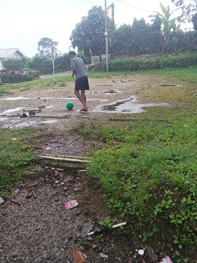 A young person in a patterned shirt and shorts plays with a green ball in a muddy, wet field. The ground is uneven and puddled. Vegetation grows sparsely. There is trash and debris visible in the foreground. In the background, a house, trees, and bushes are visible on a cloudy day.