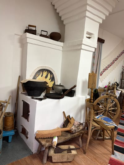 The image displays a rustic, indoor scene, likely a museum exhibit or a historical reenactment space, depicting traditional Russian village life. A large, white plaster stove dominates the center of the frame. On top of the stove, various antique household items are arranged, including two cast-iron clothes irons, a dark-colored pot, and a smaller metal pot.
In front of the stove, on a raised platform, sits a large, black cast-iron cauldron covered with a piece of fabric. Next to it are smaller metal pots and a frying pan. The front of the stove has two dark metal doors, likely for the oven and ashpit, with one visible handle. Above these doors, an arched opening in the stove is decorated with a painted depiction of flames.
To the left of the stove, several wooden objects are leaning against the wall, including long poles and a cylindrical wooden container with a lid, resting on a small blue stool.
To the right of the stove, a rustic wooden spinning wheel with a large wheel is prominently featured. It is placed behind a wooden chair with a woven mat on its seat. A stack of wooden tools and materials, including a flax hackle and a basket containing yarn, rests on a low wooden bench in front of the chair. Below the bench, a wooden trough holds more tools, including a long-handled shovel.
The floor is covered with polished wood, and a colorful striped rug runs along the right side of the frame. A white embroidered cloth with red floral patterns hangs on the wall in the background. The overall ambiance is one of historical preservation and cultural display.
While the image is identified as being located in the city of Ust'-Pyzas, Russia, there are no people visible in the scene. The objects are static and arranged for display. The environment appears to be indoors, with artificial lighting.