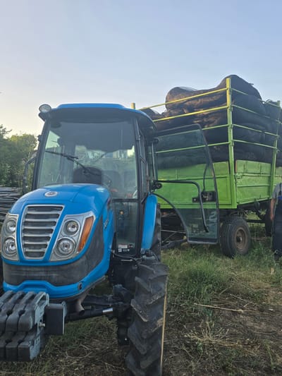 In Oreshnik, Bulgaria, a blue tractor is parked next to a green trailer overflowing with large, dark-colored sacks, likely agricultural produce. The tractor's driver-side door is open. The trailer is supported by two wheels, and a person is partially visible standing behind it. The scene is set in a grassy, outdoor environment with a clear sky.
