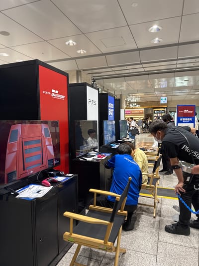 The image shows an indoor PlayStation event in Japan, possibly in Nakatsugawa. Several PlayStation 5 game stations are set up with titles like "Gran Turismo" and "Astro Bot." People are seated on director's chairs, engaging with the games. There are event staff members and signage, including "Gran Turismo" and "PlayStation" branding. A waiting line is visible in the background, along with general pedestrian traffic. The environment is a well-lit, tiled floor area, likely within a transportation hub, such as a train station or airport.