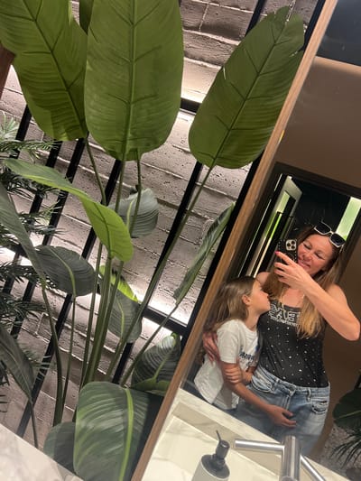 A mirror reflection shows a woman taking a selfie with a child, possibly in the bathroom of a commercial establishment in Torreforta, Spain. The woman has long hair, sunglasses on her head, and is wearing a black tank top and jeans. The child has lighter hair and is wearing a white t-shirt. A sink and soap dispenser are visible. The mirror is framed in wood and large green plant leaves partially obscure the reflection. Behind the mirror is a wall with a brick-like texture.
