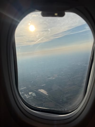 An aerial view is captured through an airplane window, looking out at a hazy landscape. The sun is visible in the upper left quadrant, casting a bright glare. The sky is a pale blue with thin, wispy clouds. Below, the ground is a patchwork of fields and structures, suggesting a rural or semi-urban area.
In Blizne Łaszczyńskiego, Poland, this image likely depicts the surrounding agricultural land and potentially the vicinity of the Blizne Łaszczyńskiego airport, as evidenced by the linear structures and uniform rectangular areas visible from above. The hazy atmosphere suggests either pollution or atmospheric conditions common at certain times of day or year. No people or specific actions are discernible from this high vantage point.