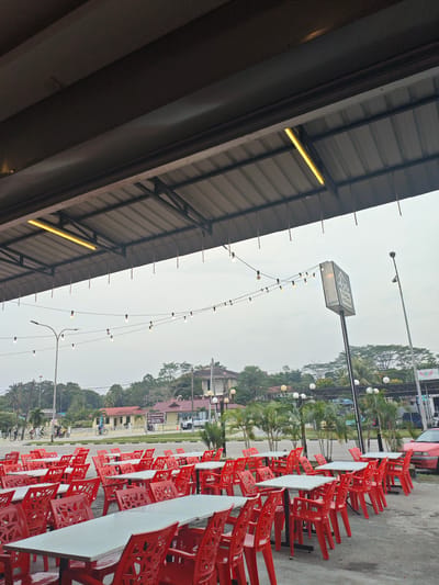 An outdoor seating area of a restaurant in Kota Tinggi, Malaysia, is shown. The area is covered by a metal roof supported by beams. String lights are hung across the open space, and a long fluorescent light is mounted on the underside of the roof. Numerous tables with white tops and red chairs are arranged neatly in rows. In the background, a street can be seen with people on bicycles and motorcycles, as well as buildings and trees. A sign for "RUMAH MAKAN" (Malay for "eatery") is visible. The sky is overcast.