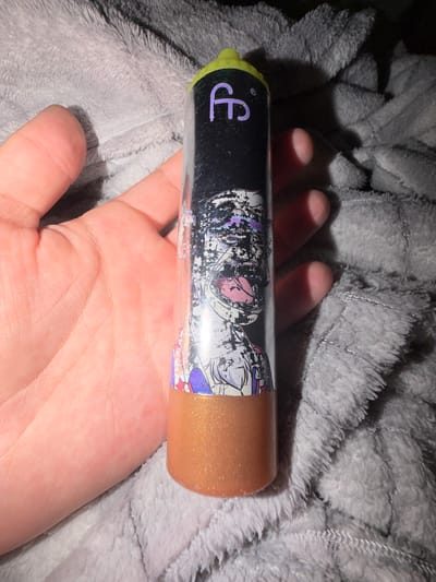 A person's hand is holding a clear cylindrical object with a lime green cap. The object is adorned with a black label featuring a purple 'ff' logo and an anime-style graphic of a character with a pink tongue. The lower portion of the object contains a shimmering golden liquid. The object is placed on a soft, grey, fuzzy textile surface. The image appears to be taken in an indoor setting, with no specific identifiable landmarks from Dorsten, Germany.