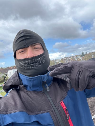 The image is a selfie of a person wearing a black balaclava with small white triangles on the face area, dark gloves, and a blue and dark gray jacket. The person is facing the camera with a slight tilt.
The background shows a cloudy sky and a residential area with houses and some trees. The location appears to be Roßdorf, Germany, based on the buildings and the general landscape visible. The photograph is taken from a high vantage point, possibly a rooftop or elevated structure.