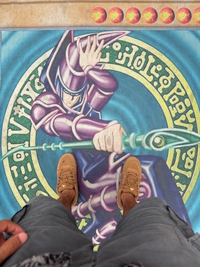 The image shows a person's legs and feet standing on a large mat featuring the Yu-Gi-Oh! card "Dark Magician." The mat is blue with a circular green and yellow design, and the Dark Magician character is depicted in purple and blue robes, holding a staff. The person is wearing grey cargo shorts and tan sneakers. The background of the image shows the top border of the mat, which has a series of small red circles with yellow arrows. The scene appears to be indoors, on a flat surface. There is no discernible El Paso, United States of America specific location information in the image.