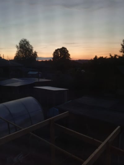 This media file captures an outdoor scene at dusk or sunset in Verkhnyaya Salda, Russia, portraying a quiet, rural or suburban landscape. The sky, which occupies the upper portion of the frame, displays a serene gradient of colors, transitioning from light blue-grey at the top to soft orange and pink hues near the horizon, typical of twilight. Subtle cloud formations are visible, suggesting clear weather.
In the midground, a dark silhouette of a tree line stretches across the horizon, punctuated by a prominent, dense tree on the left and another on the right. Below the horizon, the dark shapes of several buildings, likely houses or sheds, are visible, blending into the surrounding shadows.
The foreground features various structures in deep shadow. On the left, a translucent, arched greenhouse or tunnel-like covering is discernible. Nearby, the flat, dark roofs of what appear to be sheds or storage units are visible. A partial wooden framework, possibly part of a garden bed, a fence, or a small construction project, occupies the bottom right of the image, showing distinct horizontal and diagonal beams.
The scene is devoid of people or specific activities, conveying a sense of stillness and the peaceful end of the day. The lighting is low, emphasizing outlines and forms against the lighter sky, with most details in the lower half of the image obscured by shadow. No visible text is present.
