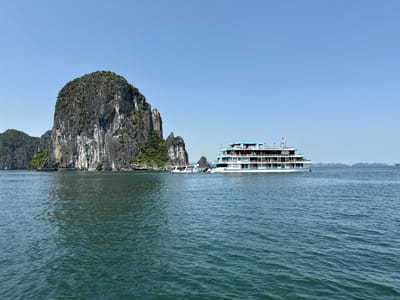 This image depicts a serene and majestic coastal landscape in Tuan Chau Ward, Vietnam, showcasing the distinctive limestone karst formations characteristic of Ha Long Bay. The scene unfolds on a clear, sunny day, with a bright, cloudless blue sky stretching across the upper portion of the frame.

The foreground and midground are dominated by the calm, dark blue-green waters of the bay, showing gentle ripples. On the left side, a massive, towering karst island rises dramatically from the water, its steep, grey rock faces textured with vertical striations and topped with dense, dark green vegetation. Other smaller karsts and islands are visible further in the distance, fading into a light atmospheric haze.

A prominent subject in the midground is a large, multi-deck cruise ship. The vessel is predominantly white with light blue accents on its upper decks and railings, featuring numerous windows and balconies. A small red flag flies from its top deck. Docked closely alongside the larger ship is a smaller, white motorboat, likely used for transfers. The registration number "QN-9953" is clearly visible on the hull of the cruise ship. On a rock formation further in the background, a yellow diamond-shaped sign with black markings is visible, possibly a navigational aid. The overall impression is one of peaceful exploration amidst stunning natural beauty.
