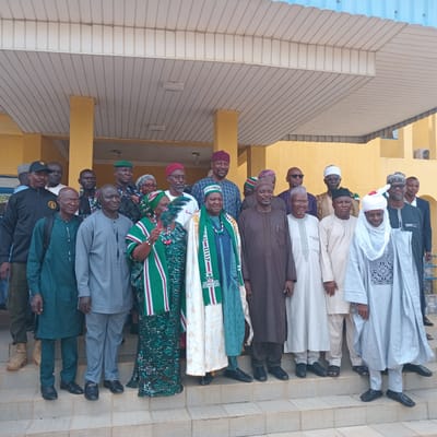 In Unguwar Gaya, Nigeria, a group of approximately twenty individuals, predominantly men with a few women, are gathered outdoors on a set of concrete stairs. They are posing for a photograph in front of a yellow building with a light blue corrugated metal roof overhead, supported by yellow pillars. The individuals are dressed in a variety of traditional and modern attire, including dashikis, caps, turbans, and some appear to be in military or security uniforms. Some individuals are wearing sashes or scarves, and one woman is holding a fan. The overall atmosphere appears formal, possibly a political or community gathering. The lighting suggests it is daytime, and the scene is viewed from a slightly low angle, looking up at the group.