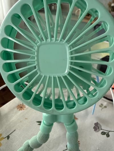 The image shows a close-up, overhead view of a teal-colored portable fan with a flexible, fuzzy green stand. The fan's protective grill is visible, with multiple spokes radiating from the center. The fan is positioned on a white tablecloth with a floral pattern, featuring small brown and yellow flowers. The background is blurred and appears to be an indoor setting. There are no people or animals visible in the image, and no specific details suggest a location within the city of Nerudstal, Ukraine. The scene is static and shows no action or interaction.
