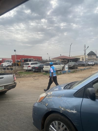 The image shows an overcast street scene in Pakuro, Nigeria. A man wearing a blue and white vest is bent over the front of a blue car with chipped paint. Behind the car, a line of parked vehicles extends into the distance, including cars, vans, and trucks. Buildings and utility poles are visible along the roadside. The sky is filled with grey clouds.