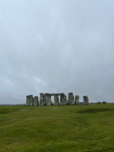 The image displays the prehistoric monument of Stonehenge, located in Larkhill, United Kingdom. The structure consists of multiple large, weathered grey sarsen stones, some standing vertically with horizontal lintels, forming a partial circular arrangement on an expansive, undulating green grass field. The sky above is uniformly overcast and light grey. In the far-right background, a group of approximately 5-7 indistinct human figures are observed standing on the grass, seemingly observing the monument. Distant vegetation or tree lines are visible along the horizon on the left side of the scene.