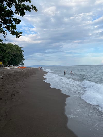 A wide-angle, vertical shot captures a tranquil beach scene in Dauin, Philippines, under a cloudy sky. The dark, volcanic sand of the beach stretches into the distance, marked by footprints and some debris. Several boats are pulled ashore near a cluster of trees on the left side of the frame. In the water, a few individuals are wading and swimming, while gentle waves break on the shore. The horizon is visible in the background, with a distant landmass under the vast expanse of clouds.