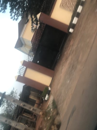 The image displays the exterior of a building, likely a multi-story residence or commercial property, captured during the daytime under what appears to be an overcast or bright sky. The building features cream-colored walls contrasted with dark brown vertical architectural accents or columns. A large, dark-colored gate, possibly made of metal or wood and appearing to be an entrance for vehicles, is centrally located on the building's facade. Above this gate, a distinct horizontal band or low wall is adorned with alternating black and white vertical stripes.
To the left of the gate, a textured, light brown panel is integrated into the cream wall. Below the gate area, a dark railing is visible, suggesting a balcony or a protective barrier. On the lower section of the building, towards the left, an outdoor air conditioning unit is mounted on the wall. The roof, partially visible on the bottom left, appears to be dark-colored, possibly grey or brown.
In the foreground, a paved surface, resembling a road or driveway, extends across the top portion of the image. Along the base of the striped wall, some green grass or low vegetation can be seen. A prominent tree with dense, dark green foliage occupies the upper left side of the foreground. Further into the background, on the right side of the image, additional sparse trees and what appear to be utility poles or vertical posts are visible. The sky, seen at the bottom of the image, is bright, suggesting the photograph was taken during daylight hours. The image appears slightly blurry, possibly due to motion or a shallow depth of field. There is no discernible activity taking place, and no text is visible in the scene.