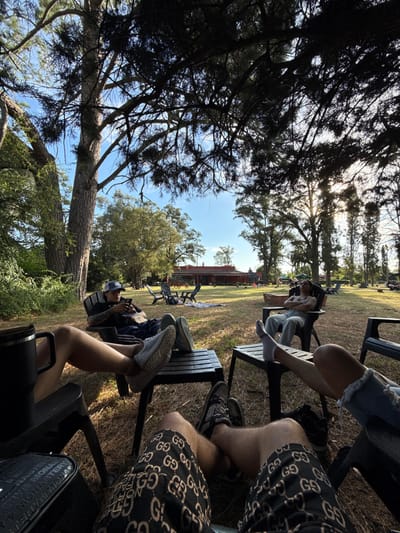 People relax on chairs in a grassy area under trees. A mix of shorts, jeans, and casual pants is visible. Some of the group are on their phones while others simply rest. A building is visible in the background, surrounded by trees. The sky is partially obscured by foliage. Chairs are uniformly black plastic. Overcast lighting creates a relaxed atmosphere.