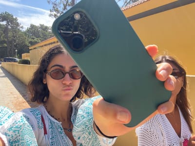 Two young women are taking a close-up selfie outdoors on a bright, sunny day in Caminha, Portugal. The primary subject on the left, with dark, wavy hair and oval sunglasses, is wearing a white and teal patterned top with multiple necklaces and is making a "duck face" expression. Her left hand is visible, partially holding the dark green-cased smartphone with a triple camera array. The second subject, partially visible on the right, has long dark hair, sunglasses, and a white crocheted top, with her right hand also grasping the phone. In the background, a long, low, yellow-painted building with a red-tiled roof is visible, framed by abundant green pine trees. A paved path runs along the left side, where a dark vehicle is partially visible in the distance under a clear blue sky.