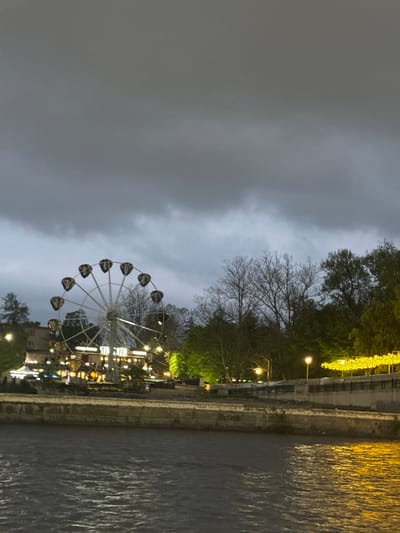 The image depicts a Ferris wheel against a cloudy sky, likely at dusk or night. The wheel is illuminated, and its carriages appear dark. Alongside the Ferris wheel, there is a river or body of water, with light reflecting off the surface, suggesting illumination from nearby structures. The opposite bank is lined with trees and buildings. Lights are strung along the edge of the river, and the scene seems to be in an urban environment. Although the city name Komgaron and its location in Russia is mentioned, there's nothing in the image to explicitly confirm this specific location.