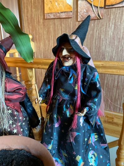 This is a close-up, eye-level shot of two decorative witch dolls, possibly intended for Halloween. The doll in the foreground is wearing a black dress adorned with colorful, holographic prints of witches, broomsticks, and crescent moons. She has bright pink hair, a black pointed hat, and a string of pearls around her neck. She is holding a small wooden broomstick in her right hand. To her left, another witch doll is visible, dressed in a shimmering red and black outfit with star patterns. The dolls are positioned in an indoor setting, with a wooden railing and wall visible in the background. The lighting is warm and natural, suggesting it may be daytime. The scene is focused on the dolls themselves, with a portion of a terra cotta pot containing dark soil in the lower left corner of the frame. The location of Габишево, Russia is not evident from the image.