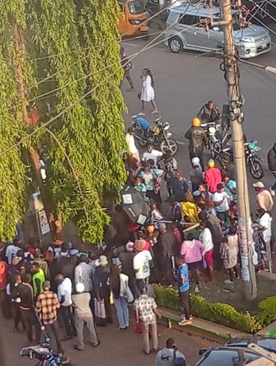 An elevated view captures a busy street scene in Murang'a, Kenya, during daylight hours. The environment features a paved road, a dense green-leafed tree dominating the left side of the frame, and a prominent utility pole with numerous overhead wires on the right. A low, manicured green hedge or planter borders a pedestrian area on the bottom right.

Approximately 50 to 70 individuals are present. A large crowd is concentrated in the mid-ground and foreground, gathered on what appears to be a sidewalk or roadside area, with some individuals spilling onto the paved road. People in the crowd wear various casual attire, including shirts, trousers, and dresses. A black open umbrella is visible within the denser part of the crowd. Actions within the crowd appear observational, with individuals standing close together.

Vehicular traffic includes an orange tuk-tuk on the upper left road, and a silver SUV (possibly a Toyota Ractis) further back on the upper right road. Closer to the crowd, several motorcycles (boda-bodas) are present; at least two are parked or stationary near the left edge of the crowd, and two individuals on motorcycles are positioned on the road on the right side of the crowd, one wearing a yellow helmet.

Separate from the main gathering, a person in a white dress is walking alone on the road in the upper mid-ground, moving right. Another individual in dark clothing walks near the large tree on the left. Signs are affixed to the trunk of the large tree on the left. A dark vehicle is partially visible in the bottom right corner of the frame.