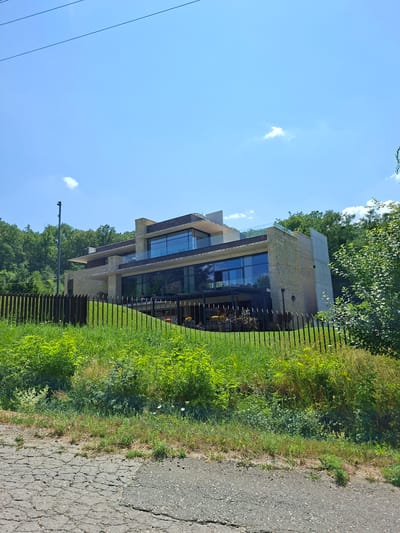 In the city of Condrița, Moldova, a modern, multi-story house is viewed from a slightly elevated position across a cracked asphalt road and a metal fence. The house, with its angular design, features stone and concrete facades, large glass windows, and cantilevered sections. A balcony with glass railings is visible on the upper level. The surrounding environment includes lush green grass and dense trees on a hillside behind the property. The sky is clear blue with a few wispy clouds. A tall pole with a security camera is positioned to the left of the house. The scene appears to be captured during daylight.